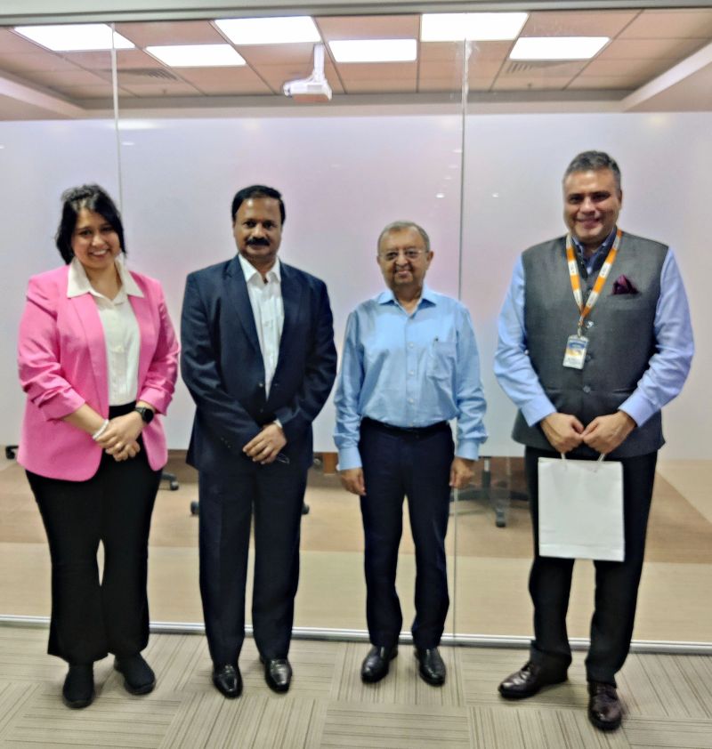 Manivannan S, second from left in a navy suit and white shirt, stands with three others in a glass-walled meeting room: a woman in a pink blazer, a man in a light blue shirt, and a man in a grey waistcoat and lanyard holding a white gift bag.